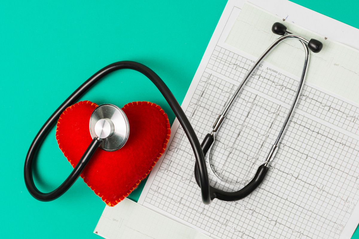 Stethoskop liegt auf einem roten Stoffherz neben einem EKG-Ausdruck auf türkisem Hintergrund